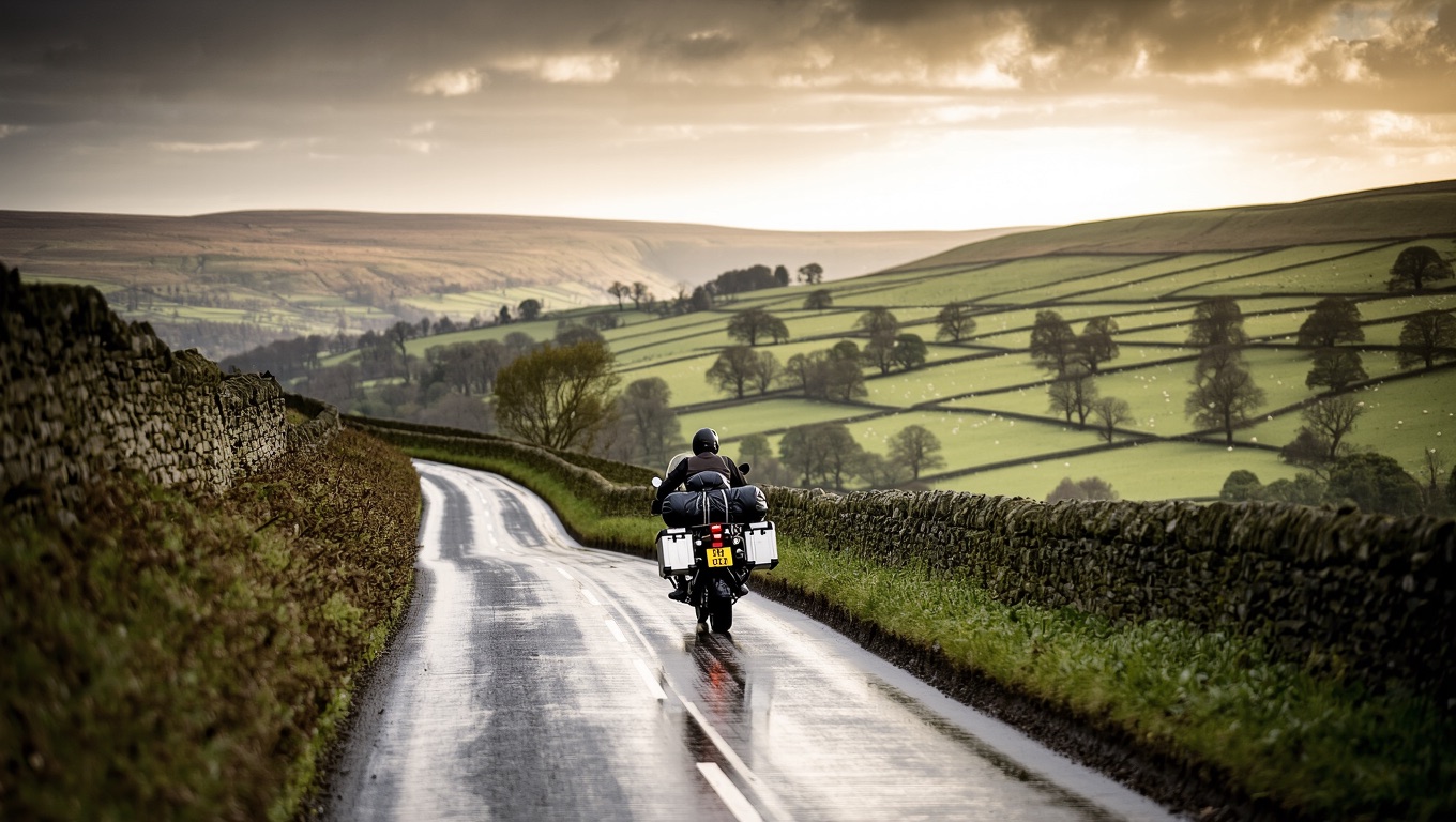 Motorcycle tourer with panniers and a roll-top drybag riding a wet single-track lane between drystone walls in the English Dales